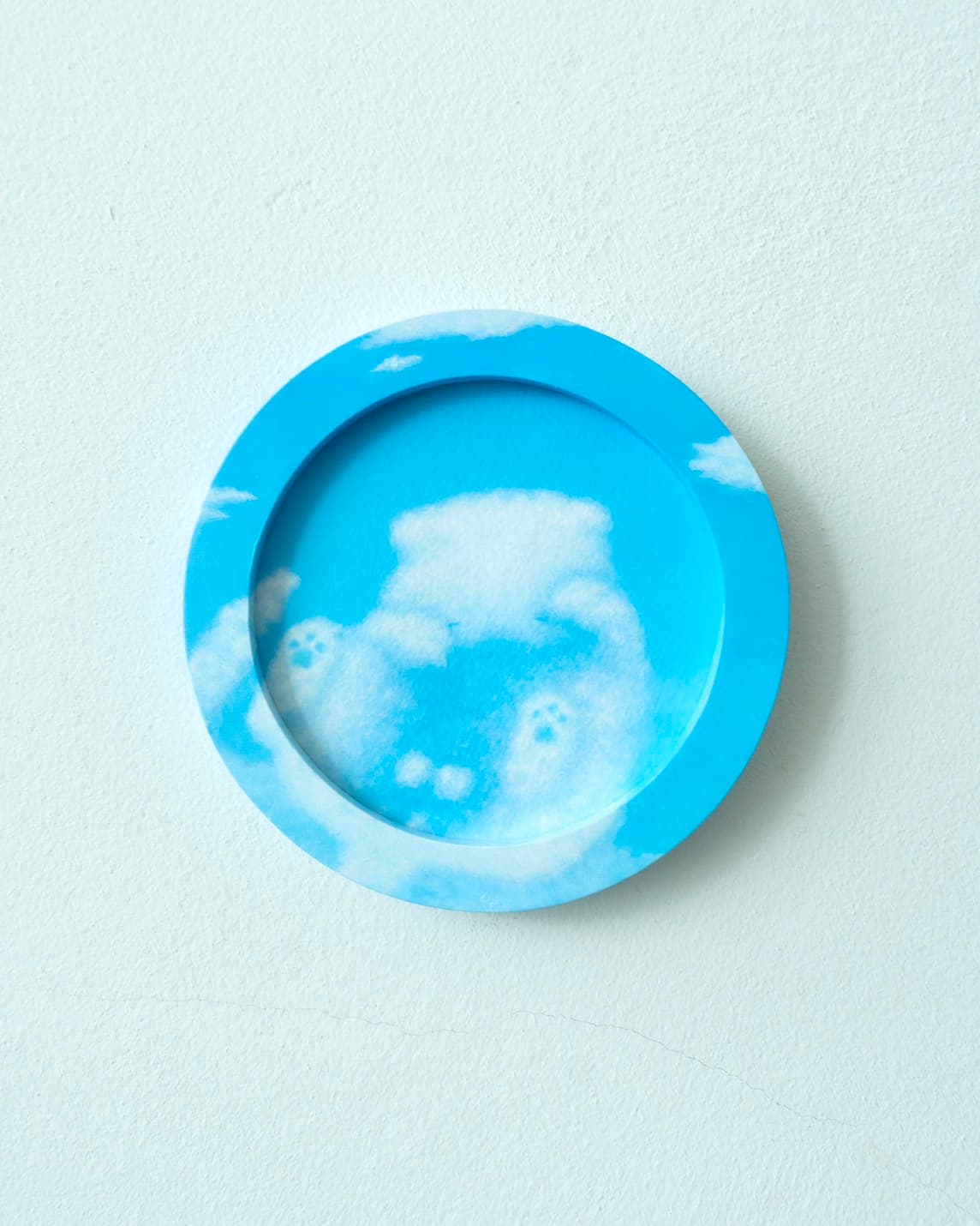 A circular painting with a sky-blue, painted rim: a cloud-cat with visible paw pads recessed at the centre